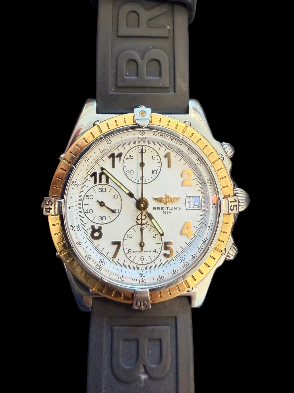 A Breitling chronograph watch with a silver-toned stainless steel case, gold-toned bezel accents, a white dial, and a dark gray silicone strap with 'BR' lettering.