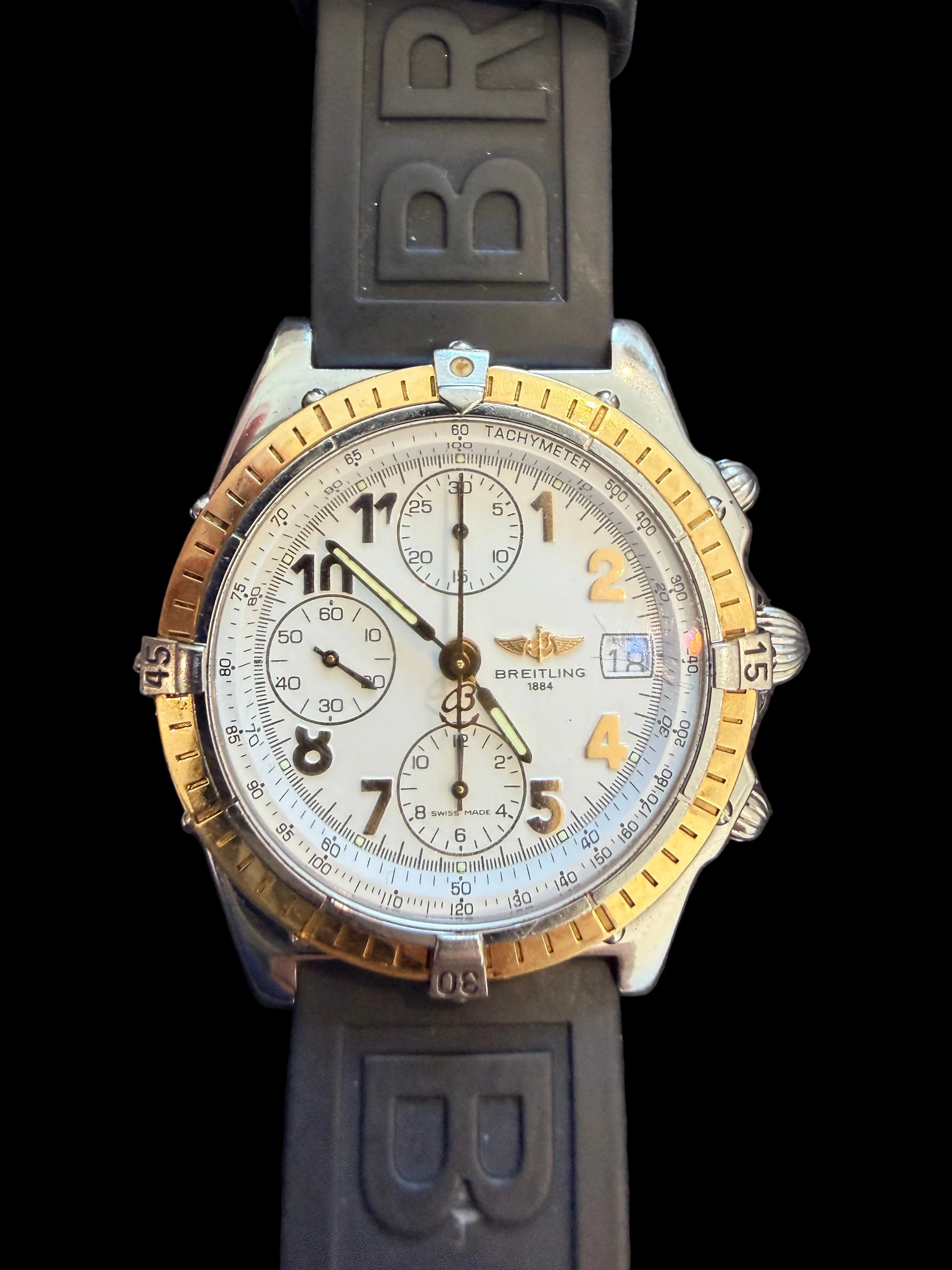 A Breitling chronograph watch with a silver-toned stainless steel case, gold-toned bezel accents, a white dial, and a dark gray silicone strap with 'BR' lettering.