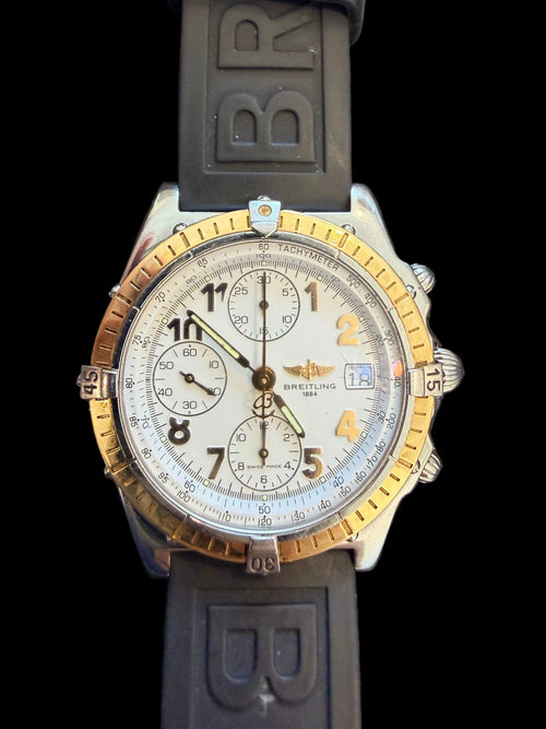 A Breitling chronograph watch with a silver-toned stainless steel case, gold-toned bezel accents, a white dial, and a dark gray silicone strap with 'BR' lettering.