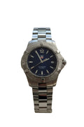 A stainless steel wrist watch with a dark blue dial and matching steel link bracelet, displayed on a beige cushion mounted on a white wall.