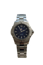 A stainless steel wrist watch with a dark blue dial and matching steel link bracelet, displayed on a beige cushion mounted on a white wall.