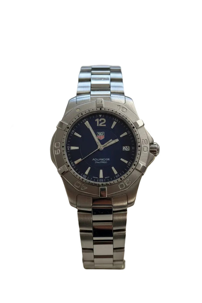 A stainless steel wrist watch with a dark blue dial and matching steel link bracelet, displayed on a beige cushion mounted on a white wall.