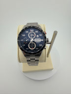 A TAG Heuer Carrera watch with a dark blue dial and a silver stainless steel bracelet, displayed on a beige cushion mounted on a white circular stand.