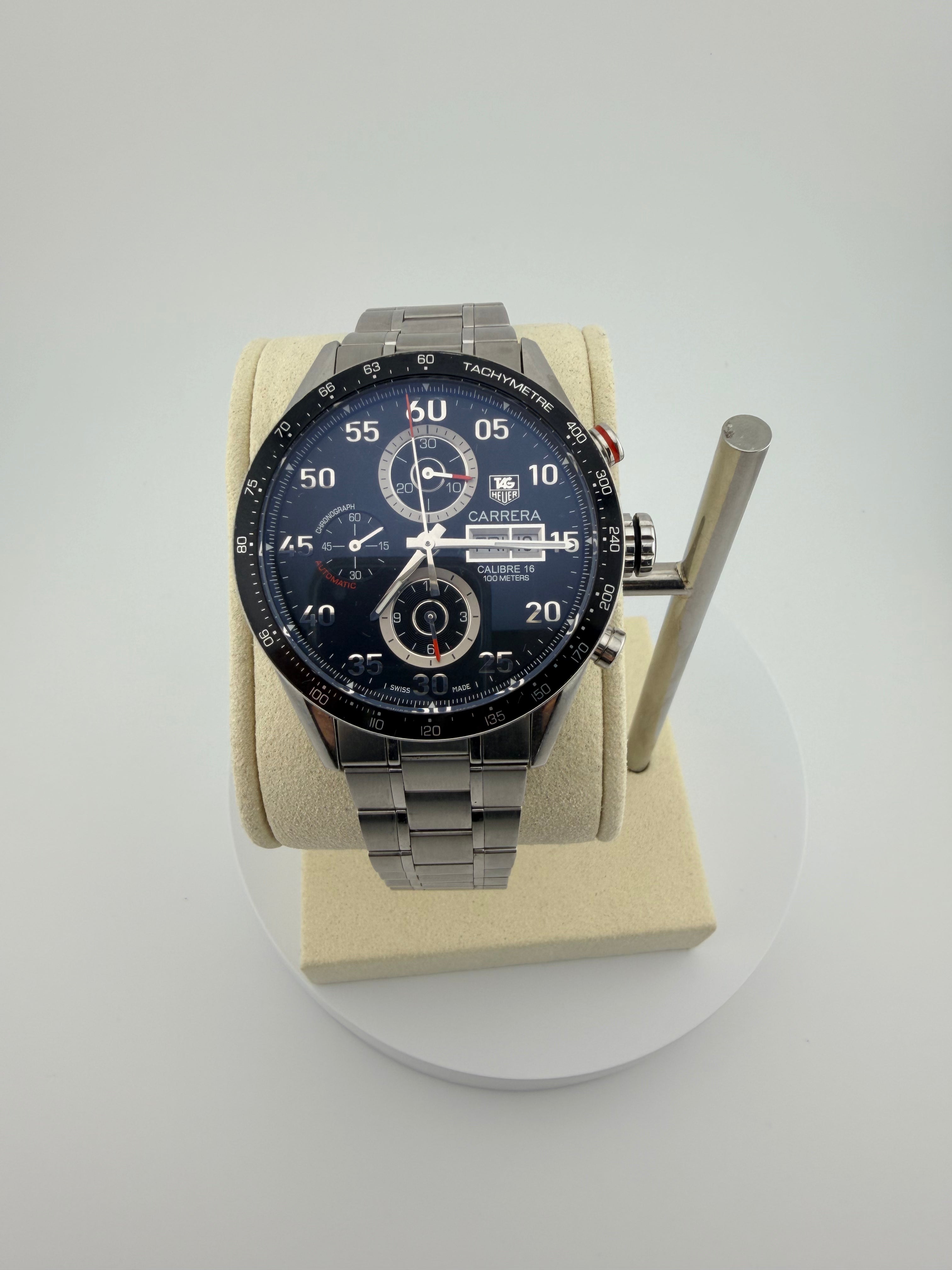 A TAG Heuer Carrera watch with a dark blue dial and a silver stainless steel bracelet, displayed on a beige cushion mounted on a white circular stand.